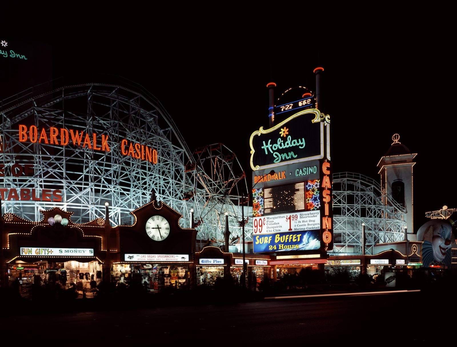 The Boardwalk casino at night: a white wooden-style roller coaster facade with red neon BOARDWALK CASINO letters, a lit Holiday Inn marquee stacked with price cards, a Ferris wheel, souvenir storefronts and a glowing clock tower against a black sky.
